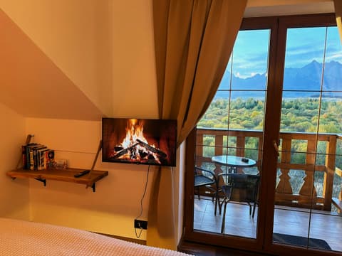 Wall TV and bookshelf under the sloping ceiling, curtains drawn back from the balcony door and the Tatras outside