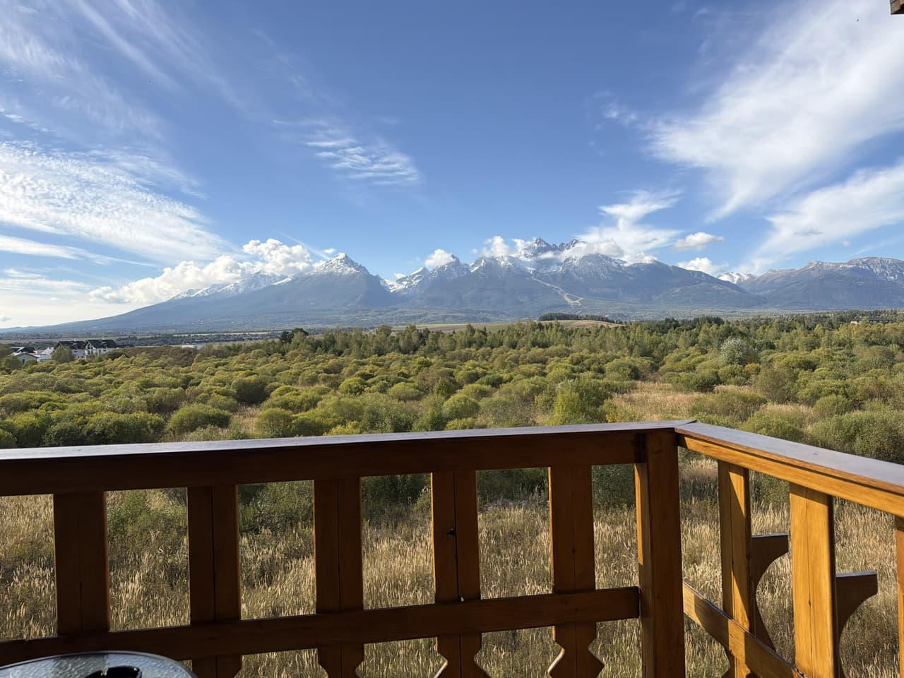 Výhľad z balkóna apartmánu – zasnežené Vysoké Tatry a Lomnický štít nad zelenou lúkou, v popredí vyrezávané drevené zábradlie.